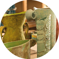 Auf dem Bild sieht man viele bunte Gef e aus Ton. Es sind zum Beispiel Vasen und Kr ge. Die Gef  e haben unterschiedliche Formen und Muster. Einige sind rund, andere sind lang oder eckig. Die Farben sind zum Beispiel gr n, t rkis, gelb, rot und grau. Manche Oberfl chen sind glatt, andere sind mit Mustern oder Strukturen verziert. Die T pferarbeiten stehen auf einem Tisch in einem hellen Raum. Im Hintergrund sieht man Regale und Kisten.