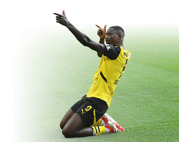 Der St rmer Serhou Guirassy von Borussia Dortmund kniet auf dem Rasen und freut sich. Er zeigt mit beiden H nden eine Jubelgeste. 