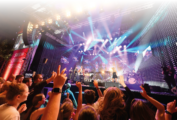 Eine jubelnde Menschenmenge steht vor einer B hne, auf der eine Band spielt. Es ist sp ter Abend und eine grelle Lichtshow erhellt die B hne. Links ist ein rotes Banner zu sehen, auf dem “SWR Sommerfestival" steht.