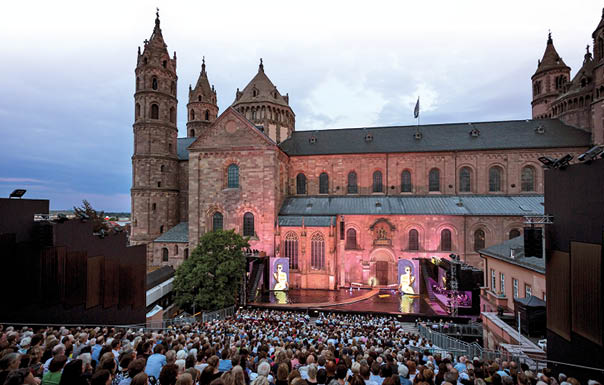 Vor der roten Sandsteinfassade des Wormser Doms ist eine Open-Air-B hne aufgebaut. Rechts von der B hne ist ein Orchester platziert. Vor der B hne steigen die Zuschauerreihen steil an. Die Reihen sind bis auf den letzten Platz gef llt.