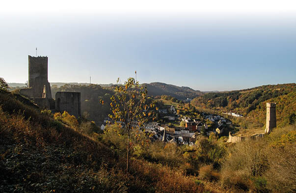 Von einem H gel blickt man ins Tal, links und rechts umrahmt von zwei Burgruinen erkennt man im Tal ein pittoreskes Dorf. Dahinter erstrecken sich am Horizont die sanften, bewaldeten H gel der Eifel.