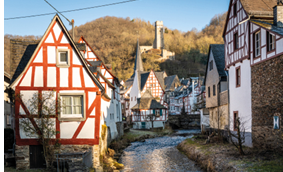 Links und rechts eines kleines Flusses stehen pittoreske Fachwerkh user. Im Hintergund erhebt sich eine Burgruine am Hang eines Berges. 