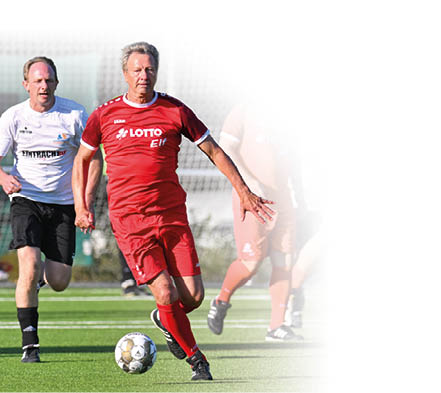 Ein Fu ballspieler in einem roten Trikot dribbelt mit einem Ball ber ein Spielfeld. Er wird von einem Spieler in einem wei en Trikot verfolgt.