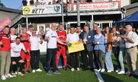 Eine Gruppe von knapp 20 Personen steht vor einem Vereinsheim aufgereiht und halten einen Symbolscheck mit LOTTO-Logo in die Kamera. Im Hintergrund sind Werbebanden und Zuschauer zu erkennen. Der Himmel ist strahlendblau.