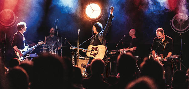 F nf ltere Rockmusiker stehen auf der B hne. Sie spielen auf ihren Instrumenten, im Hintergrund strahlen Scheinwerfer die B hne an. Im Vordergrund ist das Publikum leicht verschwommen zu erkennen.