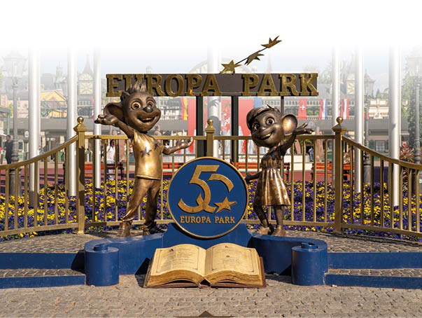 Die Euromaus und seine Freundin stehen als Bronzestatue vo einem Zaun im Europa Park. ber ihnen prangt das Europapark-Logo und in ihrer Mitte liegt ein offenes Buch, dar ber eine gro e M nze mit einer 50.