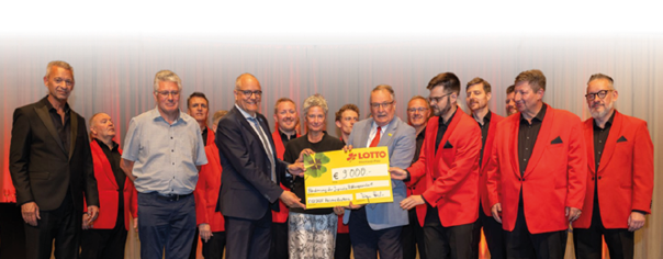 Eine Gruppe von M nnern und einer Frau steht auf einer B hne, die im Hintergrund mit einem hellen Vorhang abgehangen ist. Viele tragen Anz ge mit roten Sakkos und schwarzen Hemden. Zwei M nner und eine Frau in der Mitte halten einen gro en Spendenscheck  ber 9.000 Euro. Alle l cheln in die Kamera.