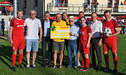 F r die bergabe des symbolischen Spendenschecks versammeln sich Spieler der Lotto-Elf, der gastgebenden Mannschaft des SV 1920 Horchheims sowie Mitarbeiter von Lotto Rheinland-Pfalz und der Lotto-Gesellschafterversammlung in einer Reihe auf dem Rasen des Sportplatzes. Der Spendenscheck wird dabei freudestrahlend in die Kamera pr sentiert.