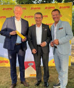 Drei M nner mittleren Alters stehen auf einer Wiese vor einer gelben Pressewand mit Lotto-Logos und l cheln in die Kamera. Der links stehende Mann h lt einen gelben Lotto-Zollstock in den H nden.