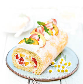 Biskuitrolle aus hellem Teig liegt auf einem hellblauen Teller. Die Rolle ist mit einer hellen Creme sowie mit Erdbeer- und Pfirsisch-St ckchen gef llt. Obenauf ist die Rolle mit Pfirsichscheiben, Erbeerh lften, Minzbl ttern und Sahne dekoriert. Neben der Rolle liegen kleine G nsebl mchen auf dem Teller.