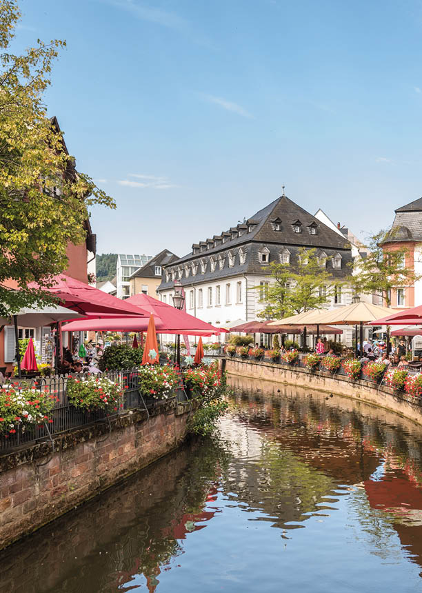 Eine Stadtansicher des Ortes Saarburg. Im Vordergrund sieht man den Leukbach. Am Ufer auf beiden Seiten sind gro e rote und gelbe Sonnenschrime z usehen. Personen sitzen an einer Au enbewirtung von Restaurants an Tischen. Bl hende Blumen am Ufergel nder und B ume runden die Szenerie an diesem sonnigen Tag mit blauem Himmel ab.