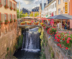 Ein kleiner Fluss mit einem Wasserfall flie t durch eine malerische Innenstadt. Auf der linken Seite steht ein Haus mit beigefarbener Hauswand und Blumenk sten an den Fenstern. Auf der rechten Seite f hrt eine Promenade entlang die am Ende mit einer kleinen Br cke ber den Fluss f hrt.  berall auf der Promenade und der Br cke stehen Sonnenschirme und Sitzgelegenheiten von kleinen Caf s. Zum Fluss hin stehtein Metallgel nder an dem Blumenk sten mit roten Geranien h ngen.