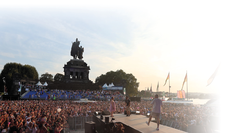 Von der B hne, auf der drei K nstler stehen, blickt man in die Zuschauermenge. Im Hintergrund ist das Kaiser-Wilhelm-Denkmal zu sehen, einige Fahnen, VIP-Zelte, mehrere B ume und die Mosel. Die Aufnahme ist in der Abendd mmerung gemacht.