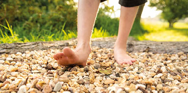 Die Nahaufnahme zeigt zwei nackte F e, die in der Natur auf  beigefarbenen Steinen gehen. Im Hintergrund sind Gr ser und Hecken zu sehen.