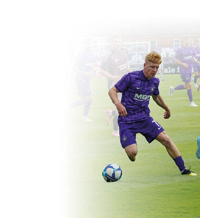 Ein blonder Fu ballspieler in einem lila Trikot l uft auf dem Fu ballplatz w hrend eines Spiels und holt mit dem rechten Bein zum Schuss auf den Ball aus, der sich kurz vor ihm befindet.