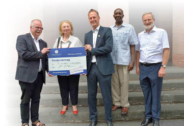 Vier M nner und eine Frau stehen auf einer Treppe vor einer Kirche und halten den symbloschen F rdervertrag von 30.000 Euro l chelnd in die Kamera.