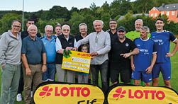 Eine Gruppe von 13 M nnern, ein Teil davon in Freizeitkleidung, der andere Teil in Fu balltrikots, steht in zwei Reihen auf dem Rasen eines lokalen Fu ballplatzes. Zwei von ihnen, die in der Mitte der Gruppe stehen, halten einen gelben Spendenscheck ber 60.500 Euro in den H nden, hinter dem noch ein kleines M dchen steht, das dar ber hervorlugt. Im Hintergrund befinden sich viele gr ne Laubb ume, der Himmel ist hellblau und klar. Vor der Gruppe auf dem Rasen stehen zwei gelbe Aufsteller mit dem Logo der Lotto-Elf.