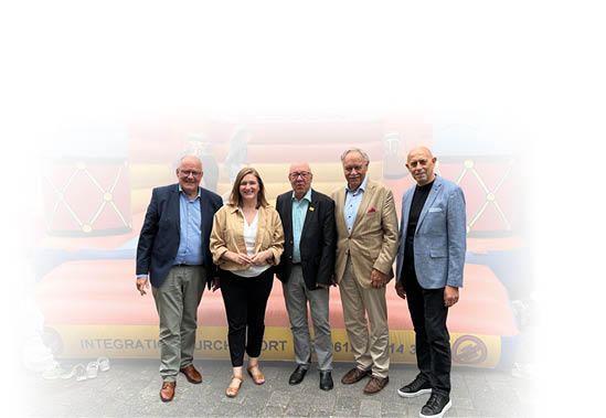 Die vier Herren tragen zwei dunkle, ein beiges und ein hellblaues Sakkos sowie drei beige sowie eine dunkle Hose. Die Ministerin steht als 2.von links und einen beigen Blazer  ber einem wei en T-Short sowie eine dunkle Hose an. Alle schauen freundlich in die Kamera.