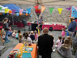 Ein Zauberer mit einer Krone aus kleinen Luftballons auf dem Kopf zaubert vor einer alten Burgmauer f r zahlreiche Kinder, die man im Vordergrund von hinten sieht. Bunte Girlanden und zwei blaue und wei e Sonnenschirme bestimmen die farbenfrohe Szenerie. Im Vordegrund steht neben einem Tisch mit orangerner Tischdecke und zahlreichen Utensilien darauf ein lterer Herr mit schwarzem Anzug.