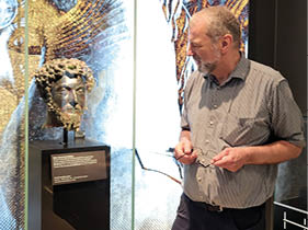 Ein lterer Mann mit kurz geschnittenem grauem Haar und Bart steht in einem Museum vor einer Glasvitrine. Er tr gt ein kurz rmliges, grau-wei  gestreiftes Hemd und eine dunkle Hose. In seinen H nden h lt er eine Brille. Sein Blick ist auf eine antike Bronzeb ste in der Vitrine gerichtet. Die B ste zeigt den Kopf eines Mannes mit lockigem Haar, vollem Bart und ernstem Gesichtsausdruck. Sie ist auf einem schwarzen Sockel montiert, vor dem eine Informationstafel angebracht ist. Im Hintergrund ist eine gro fl chige Wandgrafik in Gold- und Schwarzt nen zu sehen, die stilisierte Figuren oder Muster zeigt. Rechts im Bild befindet sich eine weitere Vitrine mit einem hellen, beschrifteten Steinfragment. Die Atmosph re ist ruhig, der Mann wirkt nachdenklich und interessiert.