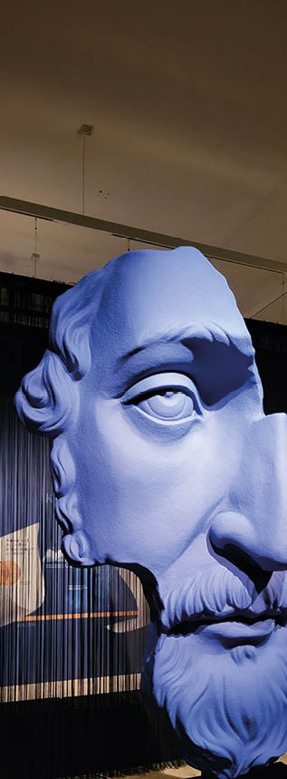 In einem abgedunkelten Museumsraum h ngt eine berlebensgro e, abstrakte Skulptur eines Gesichts von der Decke. Die Skulptur zeigt nur die linke H lfte eines m nnlichen Gesichts mit betontem Auge, Nase, Bart und lockigem Haar. Die gesamte Oberfl che ist in einem auff lligen, matten Blau eingef rbt. Der Kopf wirkt monumental und surreal. Er scheint aus vielen vertikalen, schwarzen F den zu bestehen, die wie ein Vorhang hinter und unter der Skulptur h ngen. Im Hintergrund erkennt man eine beschriftete Tafel an der Wand sowie eine leuchtende Wandgrafik in gedeckten Farben. Die Atmosph re des Raumes ist ruhig und modern, mit gezielter Beleuchtung, die die Skulptur betont.