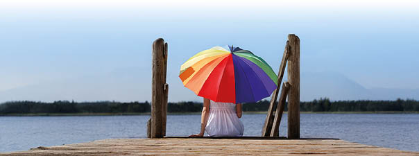 Eine Frau sitzt mittig im Bild, sie tr gt ein wei es Kleid und h lt einen bunten Sonnenschirm. Sie sitzt mit dem R cken zu uns auf einem h lzernen Steg und blickt hinaus aufs Meer, welches man im Hintergrund erkennen kann. 
