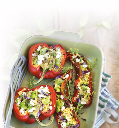 Jeweils eine rote Paprika sowie eine rote Spitzpaprika liegen der L nge nach aufgeschnitten in einer hellen Auflaufform. Gef llt sind sie mit einer Mischung aus Bohnen, Reis und Couscous. berbacken mit Feta und mit wilden Essbl ten garniert. In der Auflaufform befindet sich zudem ein grauer Pfannenwender. Die Form steht auf einem t rkis-wei  gestreiften K chenhandtuch.
