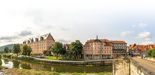 Im Vordergrund flie t ein leicht gebogener Fluss. Rechts f hrt eine Br cke ber den Fluss in die Hintergrund zu sehene Satdt hinein. De Stadt besteht auf der linken Seite aus gro en H usern mit rosa Hausw nden und rot gedeckten D chern und auf der rechten aus kleinen Fachwerkh usern. Zum Fluss hin stehen immer wieder B ume.