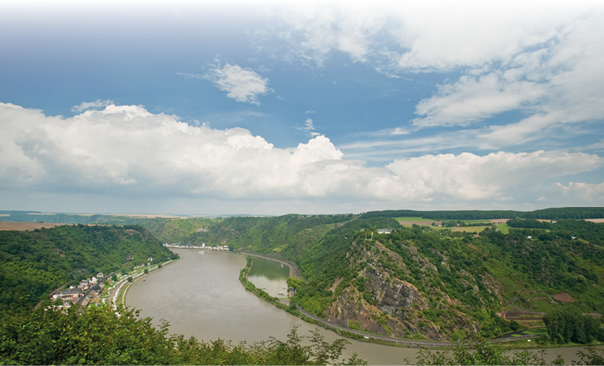 Das Foto zeigt eine weitl ufige Flusslandschaft mit dramatischer Topografie, aufgenommen von einem erh hten Aussichtspunkt namens Maria Ruh im Mittelrheintal, Deutschland. Im Mittelpunkt des Bildes schl ngelt sich der Rhein in einer markanten Kurve von links nach rechts durch das gr n bewaldete Tal. Der Fluss wirkt tr ge und breit, mit leicht br unlichem Wasser. Die Ufer sind ges umt von schmalen Stra en und kleinen Siedlungen.   Rechts im Bild erhebt sich der steile, felsige Loreleyfelsen – eine bekannte Sehensw rdigkeit. Der Felsen ist bewachsen mit B schen, B umen und Weinreben, dennoch sind die schroffen, graubraunen Felsw nde deutlich sichtbar. Oberhalb des Felsens sieht man eine Aussichtsplattform mit einem kleinen wei en Denkmal oder Kreuz.    Links am Flussufer erkennt man eine kleine Ortschaft mit wei en H usern und roten D chern, die sich entlang der Uferstra e ziehen. Auf der linken Bildseite ist das Tal etwas weiter, mit Feldern und sanfteren H geln im Hintergrund. Weiter hinten am Horizont verschmelzen gr ne H gel und Felder mit dem leicht dunstigen Himmel.    Der Himmel nimmt rund die obere H lfte des Bildes ein. Er ist  berwiegend blau mit einigen gro en, wei en, bauschigen Wolken. Die Lichtverh ltnisse sind freundlich, mit leichtem Sonnenschein, aber ohne harte Schatten – typisch f r einen leicht bew lkten Sommertag.    Im Vordergrund ist am unteren Bildrand dichter gr ner Strauchbewuchs zu sehen, der den Blick auf den Rhein leicht einrahmt.