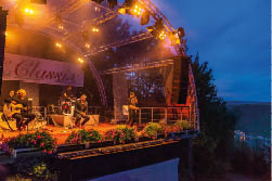 Das Foto zeigt eine stimmungsvoll beleuchtete Open-Air-B hne am Abend w hrend eines Live-Konzerts. Die B hne befindet sich im Freien auf einer Anh he mit Blick ber ein bewaldetes Tal.    Die B hne ist mit einem halbtransparenten Kuppeldach  berspannt, das von einem Ger st aus silbernen Metalltr gern getragen wird. Sie ist in warmes Licht aus orangefarbenen und gelben Scheinwerfern getaucht, die f r eine gem tliche und festliche Atmosph re sorgen. Das Licht reflektiert in der durchsichtigen Dachkonstruktion, wodurch ein stimmungsvoller Effekt entsteht.    Am vorderen B hnenrand sind Blumenk sten mit  ppigen pinken und violetten Geranien angebracht, die die B hne lebendig wirken lassen. Im Hintergrund h ngt ein gro es Banner mit der Aufschrift „Maria Ruh Classix“, was auf eine Konzertreihe oder Veranstaltungsreihe hinweist.    Auf der B hne selbst befinden sich mehrere Musiker:    Ganz links sitzt ein Gitarrist mit blonden Locken auf einem Hocker und spielt eine akustische Gitarre.    Daneben sitzt ein weiterer Mann in dunklem Anzug auf einem Hocker, offenbar ein S nger oder Moderator.    Im Hintergrund spielt ein Schlagzeuger.    Rechts steht ein weiterer S nger mit Mikrofon in der Hand und singt oder spricht gerade ins Publikum.    Die rechte B hnenseite ist leicht erh ht und mit einem gro en Lautsprecherturm best ckt. Hinter der B hne erkennt man hohe B ume, durch deren  ste das Abendlicht blitzt.    Der Himmel ist bereits tiefblau – es ist D mmerung oder kurz nach Sonnenuntergang. Am rechten Bildrand ist unterhalb der B hne in der Ferne das Rheintal zu erkennen, mit schwach leuchtenden Lichtern entlang des Flusses.