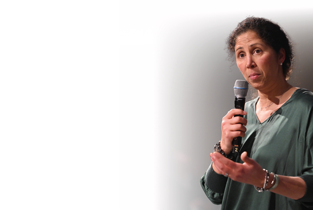 Eine Frau mittleren Alters steht in einem schwach beleuchteten Raum und spricht in ein Mikrofon. Sie hat dunkle, lockige Haare, die nach hinten gebunden sind, und tr gt eine dunkelgr ne, lang rmelige Bluse. Mit ernster und konzentrierter Miene macht sie eine erkl rende Handbewegung. Im Hintergrund sind unscharf mehrere sitzende Personen zu erkennen, die auf sie h ren. Die Atmosph re wirkt ruhig und aufmerksam, als bef nde sich die Frau in einer Vortragssituation oder Diskussionsrunde.