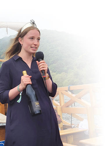 Eine junge Frau, welches ein langes, luftiges dunkelblaues Kleid tr gt. Inder linken Hand h lt sie eine schwarze Weinflasche und in der rechten ein Mikrofon, in dem sie reinredet. Auf ihren Kopf tr gt sie eine Krone. 