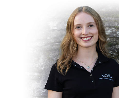 Portrait Aufnahme einer jungen Frau, sie hat schulterlange blonde Haare, welche leicht gewellt ber ihre linke Schulter fallen. Sie hat braune Augen und l chelt. Sie tr gt ein schwarzes T-Shirt auf dem „Mosel Faszination Wein“ in Versaillern steht und eine goldene Herzkette. 