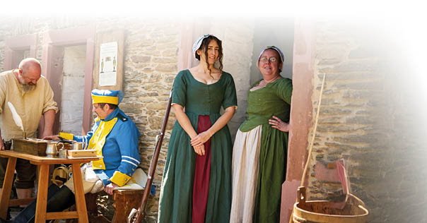 Das Bild zeigt eine Szene im Freien vor einem Geb ude aus Naturstein mit roten T rrahmen. Es sind sechs Personen zu sehen, alle in historischer Kleidung, die auf eine vergangene Epoche schlie en l sst. Die Kleidung besteht aus einfachen Stoffen in nat rlichen Farben wie Braun, Gr n, Blau und Beige.   Im linken Bildbereich sitzt eine Frau auf einer Holzbank. Sie tr gt ein braunes Oberteil, ein wei es Unterkleid und einen dunklen Rock. Neben ihr steht ein b rtiger Mann mit Glatze, der ein helles Leinenhemd und eine dunkle Hose tr gt. Er beugt sich  ber einen kleinen Holztisch, auf dem Schreibutensilien liegen, darunter ein Tintenfass mit einer wei en Feder, ein Becher und weitere Gegenst nde aus Metall und Holz.    Rechts von ihm sitzt ein Mann in auff lliger blauer Uniform mit gelben Applikationen und einer passenden M tze. Er scheint in ein Gespr ch mit dem stehenden Mann vertieft zu sein.    Rechts daneben stehen zwei Frauen im T rrahmen eines Geb udes. Die Frau vorne tr gt ein langes Kleid mit gr nem Oberteil und einem rotbraunen Einsatz im Rock. Sie steht barfu  auf dem Boden und hat die H nde gefaltet. Die Frau hinter ihr tr gt ein gr nes Oberteil und eine beige gestreifte Sch rze  ber einem wei en Rock. Sie l chelt und hat eine Hand in die H fte gest tzt.    Im Vordergrund rechts steht ein h lzerner Eimer mit einem roten Tuch darin. Auf der linken Seite des Bildes sind weitere einfache Holzm bel zu sehen, darunter eine Holzliege mit gr n-wei  gestreifter Auflage. Im Hintergrund ist ein Flechtzaun zu erkennen, dahinter gr nes Blattwerk.    Die Lichtstimmung ist freundlich und hell, die Szene wirkt belebt und detailreich.