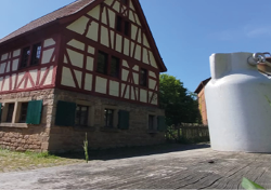 Das Bild zeigt eine l ndliche Szene bei strahlendem Sonnenschein und klarem, blauem Himmel. Im Mittelpunkt steht ein gut erhaltenes Fachwerkhaus mit auff lliger roter und wei er Balkenkonstruktion. Das Erdgeschoss besteht aus grob behauenem Naturstein, w hrend das Obergeschoss mit typischem Fachwerk gestaltet ist. Die Fensterl den im unteren Bereich des Hauses sind gr n gestrichen. Das Dach ist mit roten Ziegeln gedeckt und steil geneigt.   Links daneben ist ein weiteres Fachwerkgeb ude zu erkennen, das teilweise im Schatten liegt. Im Vordergrund erstreckt sich eine gepflasterte Fl che aus unregelm  igen Natursteinen.    Rechts im Bild, auf einem groben Holztisch oder einer Holzplattform, stehen zwei silberfarbene Milchkannen aus Metall. Die Perspektive ist leicht verzerrt durch den Weitwinkel, wodurch die Milchkannen im Verh ltnis gro  und dominant wirken. Aus einem Spalt im Holz w chst ein einzelner gr ner Grashalm hervor.    Im Hintergrund auf der rechten Seite sind weitere B ume, Wiesen mit gelben Bl ten und ein kleiner Holzzaun zu sehen, was die l ndliche und idyllische Atmosph re der Szene unterstreicht. Das Bild vermittelt Ruhe, Authentizit t und l ndliches Leben in einer vergangenen Zeit.