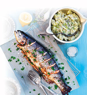 Eine gegrillte Forelle liegt eingeritzt und mit Petersilie bestreut zusammen mit einer Gabel und einem L ffel auf einem St ck hellem Backpapier auf einem Rost. Daneben steht eine wei e Sch ssel in der sich Lauchp ree beindet. Diese Sch ssel steht zusammen mit zwei Lauchzwiebeln auf einem runden Marmorbrettchen. Alles zusammen befindet sich von oben fotografiert auf einem hellblauen Tisch.