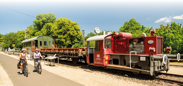 Das Bild zeigt eine sonnige Szene im Freien an einem historischen Bahnhof. Im rechten Bereich des Bildes steht eine kleine rote Diesellok mit der Fahrzeugnummer 323 108-8, die zur Deutschen Bundesbahn geh rt. Der Motorbereich der Lok ist teilweise verglast, sodass technische Bauteile sichtbar sind. Am vorderen Teil der Lok sind wei e Pufferringe und eine Trittstufe montiert. Hinter der Lok ist ein offener, rostbrauner Flachwagen sowie ein gr ner, geschlossener Waggon mit kleinen Fenstern zu sehen. Eine runde Bahnhofsuhr ist auf einem Mast auf dem Wagendach montiert.   Im linken Bildbereich verl uft ein asphaltierter Radweg parallel zu den Bahngleisen. Darauf fahren zwei Frauen nebeneinander auf Fahrr dern. Beide tragen Fahrradhelme und sportliche Kleidung in hellen Farben. Eine der Frauen hat Fahrradtaschen am Gep cktr ger befestigt. Sie fahren entspannt nebeneinander und unterhalten sich offenbar.    Der Hintergrund ist gepr gt von  ppigem, gr nem Laub und kr ftig belaubten B umen. Der Himmel ist strahlend blau mit nur wenigen kleinen wei en Wolken. Die Szenerie wirkt sommerlich und freundlich. Am linken Bildrand stehen mehrere historische Eisenbahnsignale und Informationstafeln neben dem Gleis. Der gesamte Eindruck des Bildes ist hell, klar und ruhig – es vermittelt eine harmonische Verbindung von Natur, Technikgeschichte und Freizeitaktivit t.