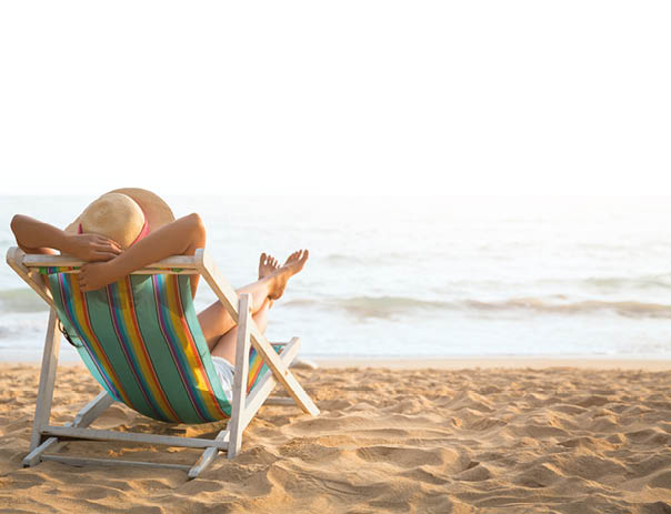 Eine Frau liegt in einem bunt gestreiften Liegestuhl an einem Sandstrand und blickt aufs Meer. Sie tr gt einen Sonnenhut mit rotem Band und hat die Arme hinter dem Kopf verschr nkt. Der Himmel ist klar und die Sonne scheint.