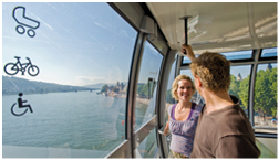 Das Bild zeigt eine Szene in einer modernen Seilbahngondel mit Panoramafenstern, die ber einen breiten Fluss f hrt.  Im rechten Bildbereich befinden sich ein Mann und eine Frau in Freizeitkleidung. Der Mann, mit kurzen braunen Haaren, tr gt ein braunes Oberteil und h lt sich an einer Stange an der Decke der Gondel fest. Die Frau, mit blonden, lockeren Haaren, l chelt ihn an und tr gt ein lila gemustertes Oberteil mit heller Hose. Beide wirken entspannt und unterhalten sich.  Im Hintergrund sieht man durch die Glasw nde der Gondel eine weite Flusslandschaft mit ruhigem Wasser. Auf der linken Flussseite erstrecken sich gr ne H gel und W lder. Rechts sind ein Uferbereich mit B umen, H usern und eine Kirche mit Turm zu erkennen – vermutlich eine kleine Stadt oder ein Dorf.  Links im Bild sind drei Symbole zu sehen, die auf Barrierefreiheit hinweisen:  – Ein Kinderwagensymbol (f r Kinderwagen geeignet)  – Ein Fahrradsymbol (Fahrradmitnahme m glich)  – Ein Rollstuhlsymbol (barrierefreier Zugang f r Rollstuhlfahrende)  Die Gondel wirkt hell, modern und gro z gig, mit einem freundlichen Ausblick auf Natur und Siedlung.  