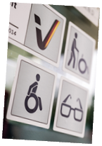 Das Bild zeigt vier quadratische Piktogramme auf einem Schild oder einer Glasfl che, die auf Barrierefreiheit hinweisen. 1. Oben links: Ein stilisiertes Symbol mit einem geschwungenen Haken, das in den Farben Schwarz, Rot und Gold gehalten ist – vermutlich ein Logo, das auf eine deutsche Institution oder ein offizielles G tesiegel hinweist.  2. Oben rechts: Eine Person, die mit einem Rollator oder einer Gehhilfe unterwegs ist. Das Symbol steht f r Barrierefreiheit f r Menschen mit eingeschr nkter Mobilit t.  3. Unten links: Ein Symbol einer Person im Rollstuhl – das international anerkannte Zeichen f r barrierefreie Zug nglichkeit f r Rollstuhlnutzer.  4. Unten rechts: Eine stilisierte Brille, die darauf hinweist, dass Angebote f r sehbehinderte oder blinde Menschen vorhanden sind.  Der Hintergrund ist leicht unscharf, was den Fokus auf die Symbole richtet. Alle Symbole sind in dunkler Farbe auf hellem Grund dargestellt und durch feine schwarze Linien voneinander abgegrenzt.  