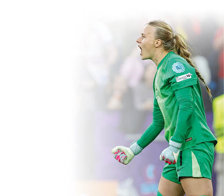 Eine Fu ballspielerin in einem gr nen Torwarttrikot jubelt lautstark. Sie steht im Profil und schaut nach links, mit weit ge ffnetem Mund und geballten F usten. Ihre langen, blonden Haare sind zu einem Zopf gebunden. Auf dem linken rmel tr gt sie ein hellblaues EM-2025-Logo. Im Hintergrund erkennt man unscharf jubelnde Zuschauer*innen, eine Person in gelber Sicherheitsweste sowie ein lila Spielfeldrand. Die Szene vermittelt gro e Emotionen und Freude – vermutlich nach einem gehaltenen Elfmeter oder einem wichtigen Sieg.