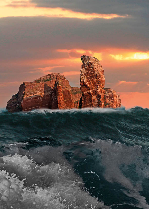 Das Bild zeigt eine dramatische Meereslandschaft bei Sonnenuntergang. Im Vordergrund tosen kr ftige, dunkelt rkisfarbene Wellen mit sch umenden Gischtkronen – das Meer wirkt aufgew hlt und kraftvoll. Im Hintergrund erhebt sich eine markante Felsformation aus dem Wasser: r tlich-braune, steil aufragende Gesteinsmassen mit klar erkennbaren Schichtlinien. Die Felsen wirken massiv und trotzen scheinbar unbeweglich den Naturgewalten um sie herum.   Am Horizont bricht die untergehende Sonne durch eine dunkle Wolkendecke und sendet warme, goldene Strahlen  ber das Wasser. Der Himmel ist in orange-, rosa- und graut nen gef rbt, was der Szenerie eine dramatische und zugleich majest tische Atmosph re verleiht.    Das Zusammenspiel aus dem wilden Meer, den imposanten Felsen und dem intensiven Licht des Sonnenuntergangs erzeugt ein kraftvolles, fast mystisches Naturbild.  