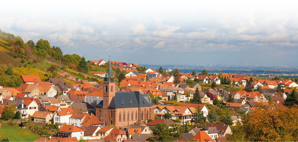 Das Bild zeigt eine idyllische Kleinstadt oder ein Dorf in einer sanft h geligen Landschaft. Im Zentrum des Ortes ragt eine gro e Kirche mit einem hohen, spitzen Turm aus rotem Sandstein hervor, deren Dach aus dunklem Schiefer gedeckt ist. Die Kirche bildet einen markanten Mittelpunkt inmitten der dicht aneinandergereihten H user mit ihren orangefarbenen, roten und braunen Ziegeld chern.   Die Ortschaft ist umgeben von Weinbergen, die sich  ber die angrenzenden H gel ziehen und durch ihre herbstliche F rbung in warmen T nen von Gelb, Orange und Rot leuchten. Zwischen den Reben, den B umen und dem gepflegten Gr n mischen sich hier und da noch Spuren des Sp tsommers.    Im Hintergrund  ffnet sich die Landschaft in Richtung eines weiten Tals, in dem sich in der Ferne eine gr  ere Stadt oder Industrieregion abzeichnet. Der Himmel ist blau und mit wei en, teils bauschigen Wolken durchzogen, was dem Bild eine freundliche, lebendige Atmosph re verleiht. Es wirkt wie eine friedliche Szene aus einem gut erhaltenen historischen Weinort.  