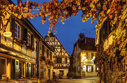 Das Bild zeigt eine enge, gepflasterte Gasse mit Fachwerkh usern. Links h ngt an einem Haus ein ovales Schild mit der Aufschrift “Friseur". rechts ist ein Geb ude mit dem Schriftzug "Weinstube". Auf den Fensterb nken stehen Blument pfe mit rosa Blumen. Der Himmel ist dunkelblau und ber der Gasse verl uft im oberen Bildbereich eine Girlande mit Bl ttern. 