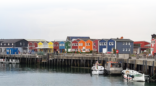 Das Bild zeigt eine Hafenpromenade mit einer Reihe bunter H user, die direkt am Wasser stehen. Die H user sind zweist ckig und in verschiedenen kr ftigen Farben gestrichen, darunter Gelb, Rot, Gr n, Orange, Blau und Grau. Vor den H usern verl uft ein breiter Holzsteg oder eine Mole, auf der einige Menschen spazieren oder sitzen. Am Wasser liegen mehrere kleine Boote und Yachten angelegt, darunter ein wei es Boot mit der Aufschrift „Helgoland“. Der Himmel ist leicht bew lkt, und das Wasser wirkt ruhig. Die Atmosph re wirkt freundlich und einladend, typisch f r einen kleinen, malerischen Hafenort.
