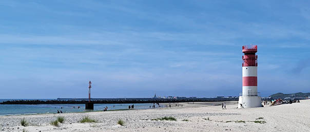 Auf dem Bild ist ein Strand mit hellem Sand zu sehen, auf dem vereinzelt kleine Grasb schel wachsen. Rechts im Bild steht ein rot-wei gestreifter Leuchtturm, der auff llig und gut sichtbar ist. Im Hintergrund verl uft eine Mole oder ein Wellenbrecher aus dunklen Steinen, der ins Meer hineinragt. Dahinter ist das Meer mit ruhigem, blauem Wasser zu erkennen, das sich bis zum Horizont erstreckt. Am Strand und im Wasser sind mehrere Menschen zu sehen, die das sch ne Wetter genie en. Der Himmel ist klar und blau, mit nur wenigen d nnen Wolken. Insgesamt vermittelt das Bild eine ruhige, sonnige Atmosph re an einem Strandtag.  