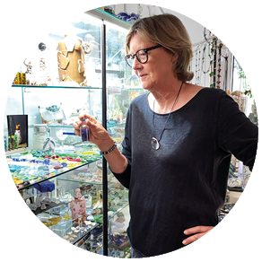 Auf dem Bild sieht man eine Frau mittleren Alters mit kurzen, hellbraunen Haaren und einer Brille. Sie tr gt ein dunkelblaues Langarmshirt und eine blaue Hose. Um den Hals hat sie eine Kette mit einem gro en Anh nger, an ihrem linken Handgelenk sind mehrere Armb nder und eine Uhr zu sehen.   Die Frau steht vor einer gro en Glasvitrine, die mit vielen bunten, kleinen Glasobjekten und verschiedenen Dekorationsgegenst nden gef llt ist. Sie h lt in ihrer rechten Hand ein kleines Glasfl schchen mit lilafarbenen K rnern oder Perlen und betrachtet es aufmerksam.    Der Raum ist hell erleuchtet, durch ein gro es Fenster im Hintergrund f llt Tageslicht herein. Im Hintergrund sind weitere Regale und Vitrinen mit vielen weiteren kleinen Objekten zu erkennen. Insgesamt wirkt die Szene wie in einem Kunsthandwerksatelier oder einem Museum f r Glasarbeiten oder Sammlerst cke.    M chtest du noch eine detailliertere Beschreibung oder Informationen zu einzelnen Objekten?  