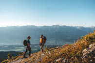 Auf dem Bild sind zwei Personen beim Wandern in den Bergen zu sehen. Beide tragen Rucks cke und Outdoor-Bekleidung, wobei der Mann links eine Kappe tr gt und der Frau rechts eine Sonnenbrille aufhat. Sie befinden sich auf einem Pfad, der von trockenem Gras und kleinen Str uchern ges umt wird. Im Hintergrund erstreckt sich ein gro es Bergpanorama mit klar erkennbarem blauen Himmel dar ber. Ganz unten im Tal sieht man eine Siedlung oder kleine Stadt. Das Licht scheint warm zu sein, wahrscheinlich kurz vor Sonnenuntergang oder nach Sonnenaufgang, was der Szene eine angenehme Atmosph re verleiht. 