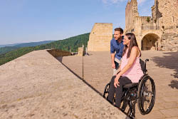 Das Bild zeigt eine Frau im Rollstuhl und einen Mann, die gemeinsam eine historische Burganlage besuchen. Die beiden befinden sich auf einem weitl ufigen Burghof aus hellem Steinpflaster, flankiert von imposanten, sandsteinfarbenen Mauern mit romanischen Fenstern und B gen. Die Burg wirkt gut erhalten und vermittelt ein Gef hl von Geschichte und Erhabenheit.   Die Frau tr gt ein rosafarbenes Oberteil und schwarze Hose, der Mann ein blaues Hemd und Jeans. Beide blicken l chelnd in die Ferne – offenbar genie en sie gemeinsam die Aussicht  ber die gr ne, bewaldete H gellandschaft, die sich weit unterhalb der Burg erstreckt. Die Szene spielt bei sonnigem Wetter, unter einem fast wolkenlosen, blauen Himmel.    Ein gro er Baum am rechten Bildrand spendet Schatten, was dem Bild eine warme, friedliche Atmosph re verleiht. Die Szene vermittelt Inklusion, Lebensfreude und Entdeckergeist in einer barrierefrei zug nglichen Kulturst tte.