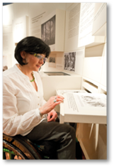 Das Bild zeigt eine Frau im Rollstuhl, die sich in einer Ausstellung oder einem Museum befindet. Sie tr gt eine wei e Bluse, eine graue Hose und auff lligen Schmuck – darunter gr ne Ohrringe, eine passende Halskette sowie Ringe und Armb nder. Die Frau hat dunkle, schulterlange Haare und eine Brille mit leicht get ntem Glas.   Sie liest aufmerksam einen Informationstext, der auf einer ausziehbaren Schublade pr sentiert wird. Der Text enth lt neben dem Flie text auch eine Illustration. Die Frau scheint interessiert und konzentriert. Die Umgebung ist modern und hell gestaltet, mit zahlreichen Schautafeln, Texten und historischen Fotografien an den W nden.    Die Szene vermittelt einen inklusiven Zugang zur Kultur: Die Ausstellung ist barrierefrei gestaltet, was durch die Zug nglichkeit der Informationen in angenehmer H he und Form deutlich wird. Die Atmosph re wirkt ruhig, lehrreich und respektvoll.  